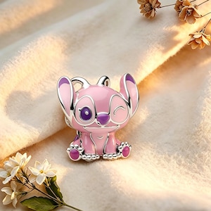 Pode incluir: Um berloque rosa e prateado com um personagem de desenho animado com orelhas grandes e uma express&atilde;o piscando. O berloque tem detalhes roxos nas orelhas, olhos e p&eacute;s. O berloque est&aacute; sobre uma superf&iacute;cie macia e esbranqui&ccedil;ada.