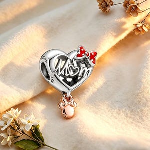 May include: A silver charm with a heart shape and the word "Mom" in cursive. A red bow with white polka dots is on top, and a rose gold bow-shaped charm hangs below. The charm is a jewelry item.