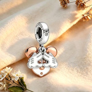 Op de afbeelding: Een zilveren en ros&eacute;gouden bedel met een Mickey Mouse-ontwerp. De bedel heeft Mickey's handen die een hartvorm vormen, met ros&eacute;gouden oren en een wit gezicht. De bedel hangt aan een zilveren beugel met het Pandora-logo.