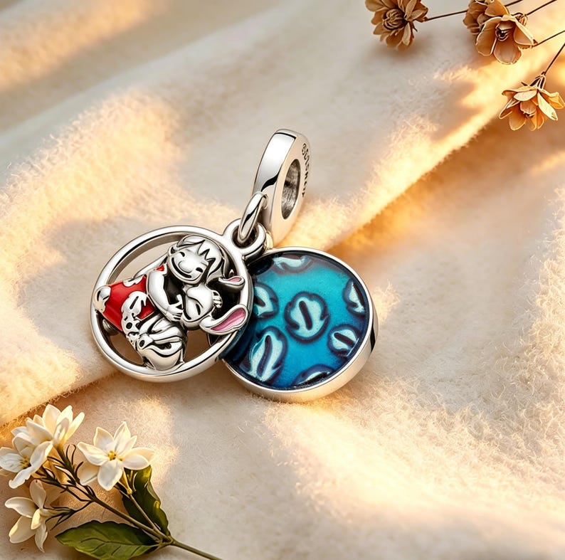 May include: A silver charm pendant with a cartoon character in a red outfit hugging a rabbit, set in a circular frame. It includes a blue enamel disc with a pattern. The charm is designed to be a pendant.
