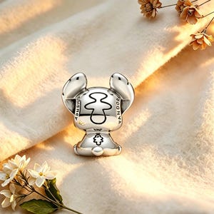 May include: A silver charm featuring a stylized character with large ears. The charm has the words "DISNEY" and "PANDORA" engraved on it. The charm is on a soft, cream-colored surface, with flowers in the background.