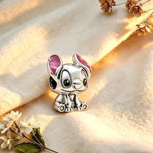 May include: A silver-colored charm in the shape of a cartoon character with large black eyes and pink-lined ears. The charm is sitting down and has a detailed design. The background is a soft, off-white fabric with flowers.