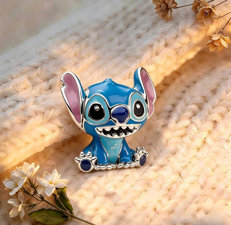 May include: A metal enamel pin featuring the character Stitch with bright blue skin, large ears with pink inner sections, and a wide grin showing white teeth. The character has large black eyes and a dark blue nose, rendered in a glossy finish.