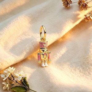 May include: A gold-colored charm in the shape of a toy soldier, with pink and white accents. The charm has a star on the hat and is suspended from a gold loop. The charm is a festive holiday-themed accessory.