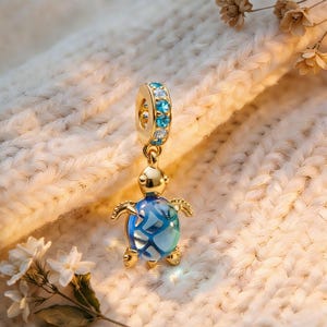 Könnte beinhalten: Ein goldfarbener Schildkröten-Anhänger mit blau-klarer Schale und Akzenten. Der Anhänger hat oben eine Schlaufe mit blauen und klaren Steinen. Der Anhänger hängt an einer weißen Strickoberfläche.