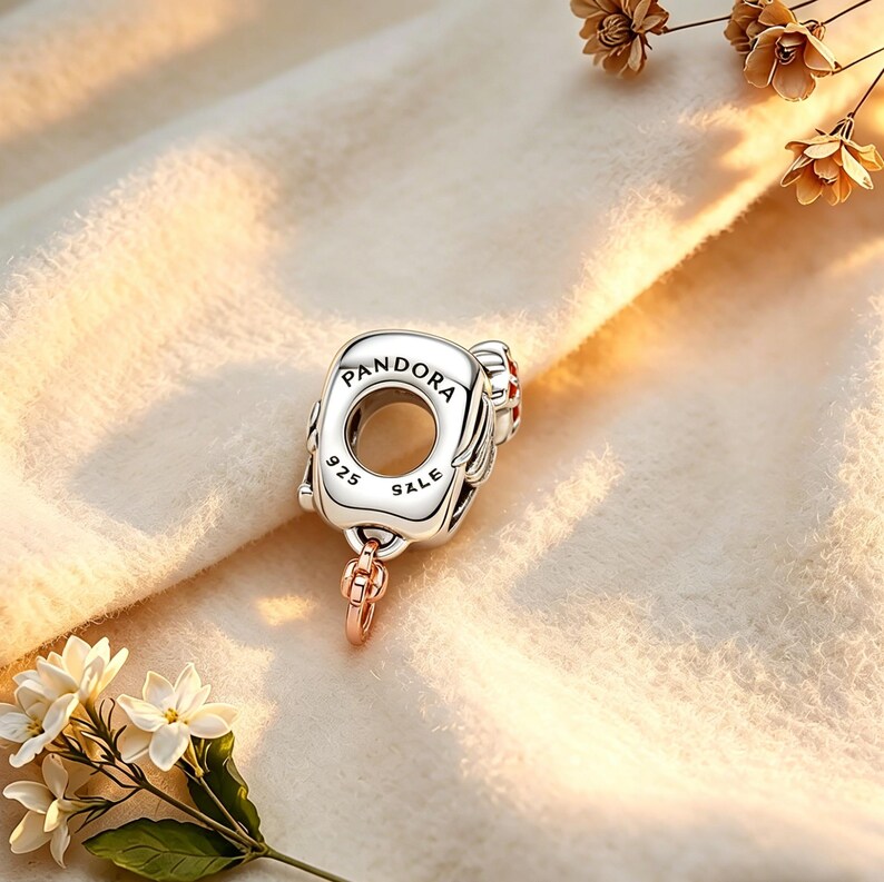 May include: A silver charm with the word "Pandora" engraved on it. The charm has a circular design with the text "925 SALE". A small rose gold chain is attached to the bottom. The charm is on a cream-colored surface.