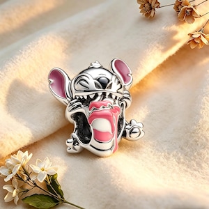 May include: A silver charm depicting the Disney character Stitch. The charm has pink detailing on the ears and tongue, with black eyes and a detailed design. The charm is set against a soft, cream-coloured background.