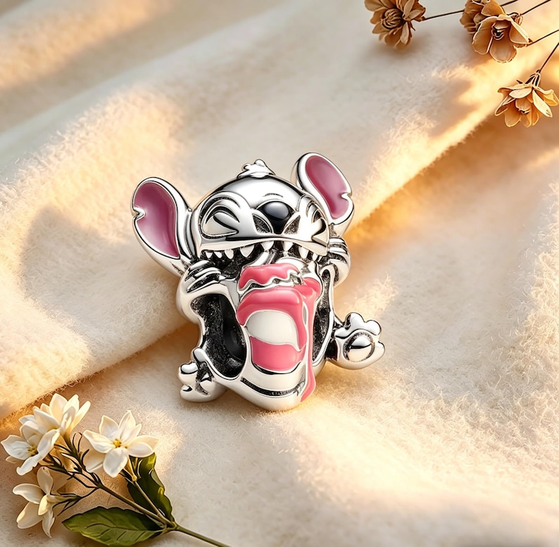 May include: A silver charm depicting the Disney character Stitch. The charm has pink detailing on the ears and tongue, with black eyes and a detailed design. The charm is set against a soft, cream-coloured background.