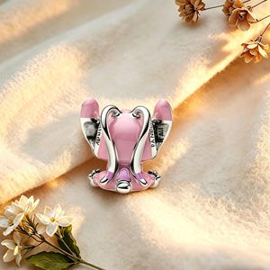 Pode incluir: Um charm rosa e prateado em forma de personagem de desenho animado, com orelhas grandes e uma postura sentada. O charm tem a palavra "PANDORA" nos detalhes prateados. Est&aacute; sobre um fundo macio, creme, com detalhes florais.
