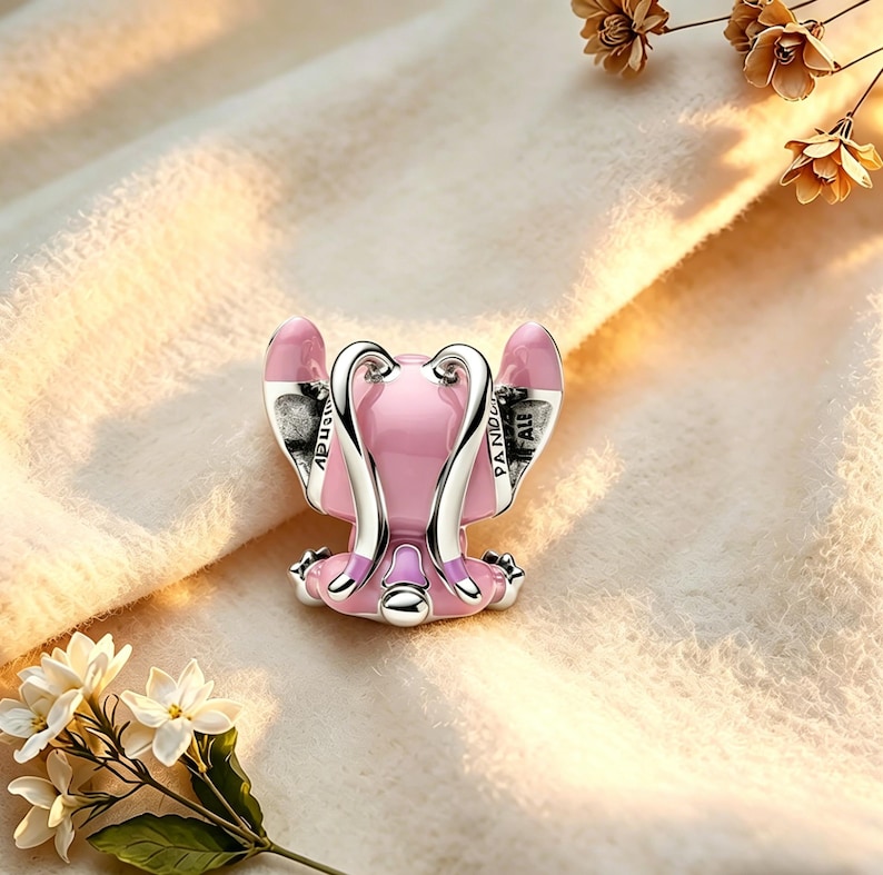 Pode incluir: Um charm rosa e prateado em forma de personagem de desenho animado, com orelhas grandes e uma postura sentada. O charm tem a palavra "PANDORA" nos detalhes prateados. Est&aacute; sobre um fundo macio, creme, com detalhes florais.