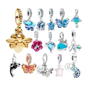 Puede incluir: Una colección de dijes de metal con varios diseños, incluyendo una luciérnaga dorada, una mariposa azul, una tortuga marina, una flor rosa, un corazón, una ballena, un delfín y un camaleón. Algunos incluyen el texto You light my way y You are magic.