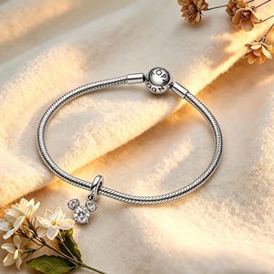 May include: A silver bracelet with a Mickey Mouse charm. The charm has a large, round, clear crystal head and two smaller crystals for ears. The bracelet is on a cream-coloured surface with flowers. The bracelet is made of silver.