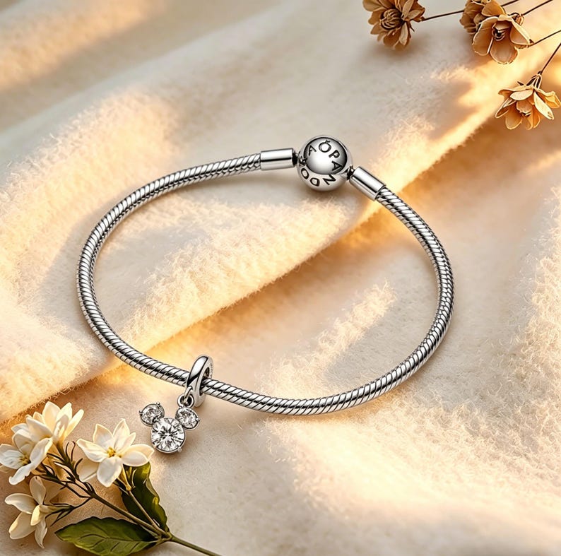 May include: A silver bracelet with a Mickey Mouse charm. The charm has a large, round, clear crystal head and two smaller crystals for ears. The bracelet is on a cream-coloured surface with flowers. The bracelet is made of silver.