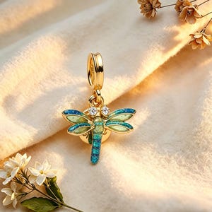 Könnte beinhalten: Ein goldfarbener Libellen-Anhänger mit blau-grünen Emailleflügeln und zwei klaren Kristallakzenten. Die Libelle hat ein detailliertes Design und ist an einer goldfarbenen Öse aufgehängt.