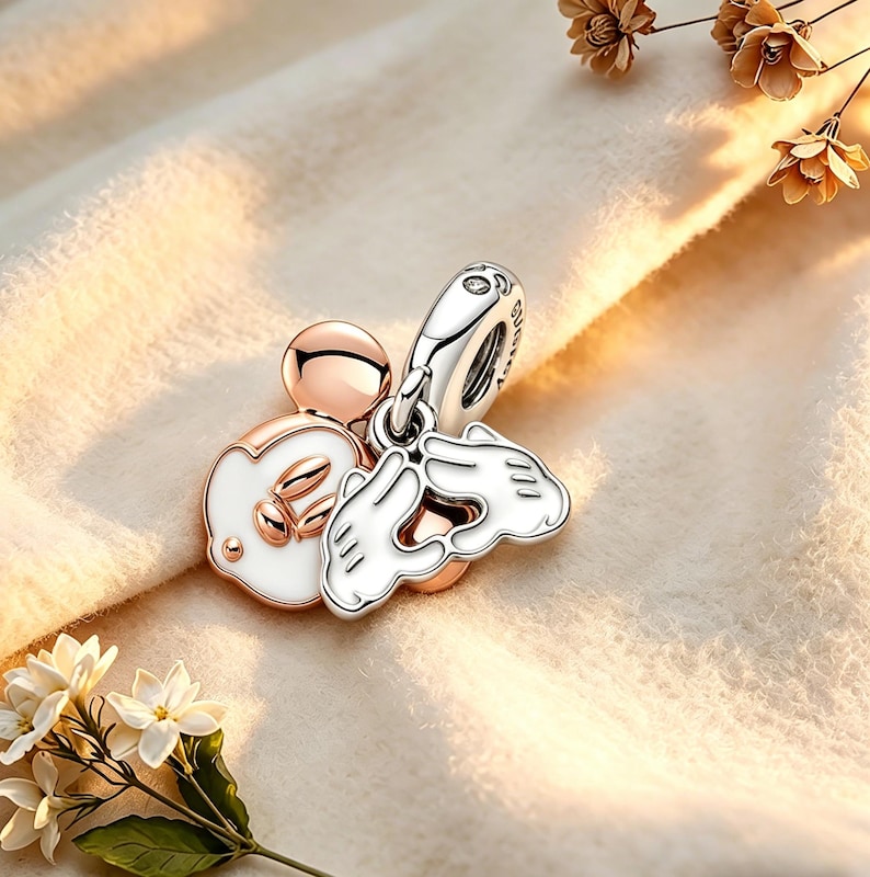 Op de afbeelding: Een bedel met een ros&eacute;gouden Mickey Mouse-hoofd en -handen, met witte emaille details. De bedel is bevestigd aan een zilveren beugel. Het ontwerp omvat een kleine, ronde ros&eacute;gouden kraal en een wit ge&euml;mailleerd Mickey Mouse-hoofd met gouden accenten.