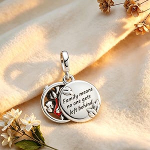 May include: A silver pendant with a hinged design. One side features a floral pattern with red accents, while the other side has the inscription "Family means no one gets left behind." The pendant is suspended from a silver loop.
