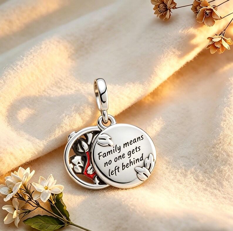 May include: A silver pendant with a hinged design. One side features a floral pattern with red accents, while the other side has the inscription "Family means no one gets left behind." The pendant is suspended from a silver loop.