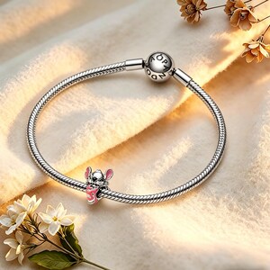 May include: A silver charm bracelet featuring a cartoon character charm. The charm is a pink and white character holding a pink object. The bracelet has a round clasp with the word "PANDORA" engraved. The bracelet is on a cream-coloured surface.