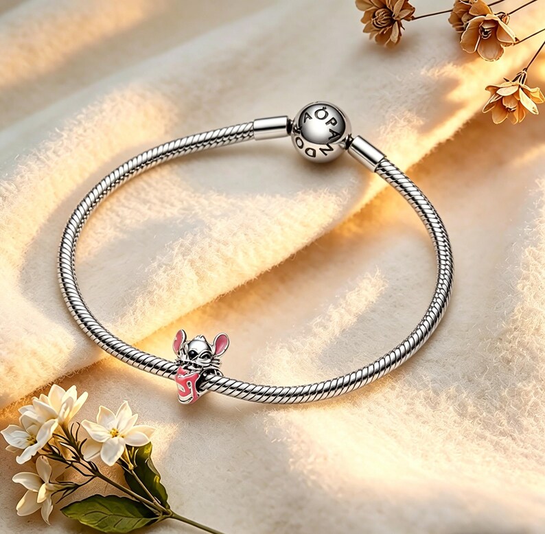 May include: A silver charm bracelet featuring a cartoon character charm. The charm is a pink and white character holding a pink object. The bracelet has a round clasp with the word "PANDORA" engraved. The bracelet is on a cream-coloured surface.