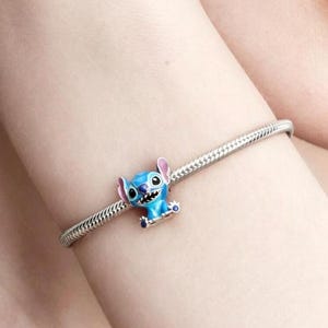May include: A silver-toned snake chain bracelet featuring a small, blue enamel charm of the character Stitch. The charm depicts the creature with large ears, a wide grin, and dark eyes, securely attached to the flexible metal link chain.