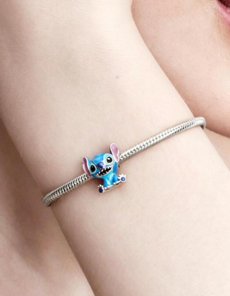 May include: A silver-toned snake chain bracelet featuring a small, blue enamel charm of the character Stitch. The charm depicts the creature with large ears, a wide grin, and dark eyes, securely attached to the flexible metal link chain.