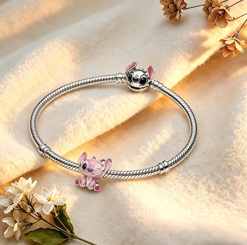 Pode incluir: Uma pulseira de prata com dois berloques com um personagem de desenho animado rosa e preto. Um berloque est&aacute; no fecho e o outro &eacute; um berloque pendurado. A pulseira est&aacute; sobre uma superf&iacute;cie branca e macia com flores.