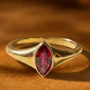 以下が含まれることがあります： マーキス型の深紅の宝石が付いたゴールドリング。リングは滑らかで磨かれたバンドと石を支えるセッティングを備えています。背景は暖かくアースカラーで、上部に「GRIFFANY ATELIER」の文字があります。