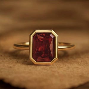 May include: A gold ring featuring a rectangular, deep red gemstone. The gemstone is set within a gold bezel, and the ring band is a simple, polished gold. The ring is displayed on a textured, brown surface.