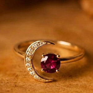 Op de afbeelding: Een gouden ring met een halve maan ontwerp, bezet met kleine, heldere stenen. De ring heeft een ronde, dieprode edelsteen. De ring wordt gepresenteerd tegen een warme, getextureerde achtergrond, wat een luxueus gevoel creëert.