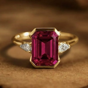 May include: A gold ring featuring a large, rectangular, deep red gemstone as the centerpiece. Two smaller, triangular, clear gemstones flank the main stone. The ring is set on a brown surface.