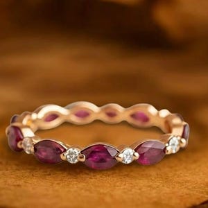 May include: A gold ring featuring alternating oval-cut rubies and small round diamonds. The ring is displayed on a brown surface. The words "GRIFFANY ATELIER" are at the top of the image.