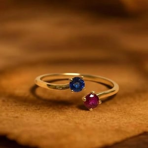 May include: A gold ring with a split design, set with a blue gemstone and a red gemstone. The ring is displayed on a brown surface. The text "GRIFFANY ATELIER" is visible at the top.