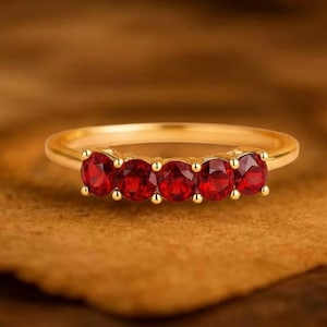 Op de afbeelding: Een gouden ring met vijf ovaal geslepen, helderrode edelstenen. De ring ligt op een warme, getextureerde ondergrond, met de inscriptie "GRIFFANY ATELIER GRIFFANY ATELIER" bovenaan. Eenvoudig en elegant ontwerp.