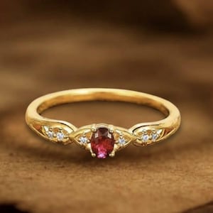 May include: A gold ring with a central oval-cut ruby and small accent diamonds. The ring has a delicate, ornate design with a polished finish. The ring is displayed on a brown surface, showcasing its elegant details.