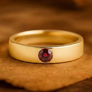 May include: A gold-colored ring with a single, round, red gemstone set in the center. The ring has a smooth, polished band. The background is a warm, brown tone, with the text "GRIFFANY ATELIER GRIFFANY ATELIER" at the top.