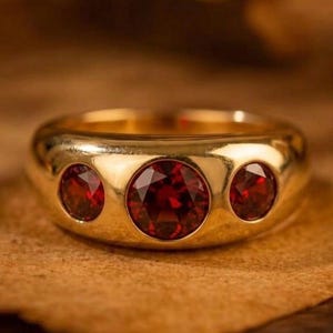 May include: A gold ring with three round, deep red gemstones. The ring has a smooth, polished surface and is set on a textured, brown surface. The text "GRIFFANY ATELIER" is visible at the top of the image.
