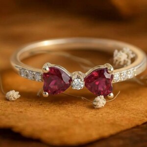 May include: A gold-toned ring featuring a bow design with two heart-shaped, deep red gemstones and a small diamond center. The ring band is encrusted with small, clear gemstones. The ring is set on a brown surface.