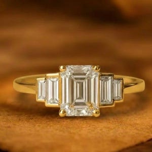 May include: A gold ring featuring an emerald-cut center stone flanked by two smaller, rectangular stones on each side. The ring is set on a gold band and is displayed on a brown surface. The text "GRIFFANY ATELIER" is at the top.