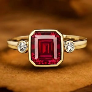 Könnte beinhalten: Ein goldener Ring mit einem großen, quadratisch geschliffenen, tiefroten Edelstein in der Mitte, der von einer goldenen Lünette umrahmt wird. Zwei kleinere, runde, klare Edelsteine ​​sind auf beiden Seiten des zentralen Steins gesetzt. Der Ring liegt auf einer braunen Oberfläche.