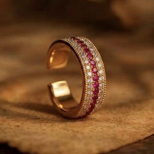 Puede incluir: Un anillo abierto de oro con una fila central de piedras circulares de color rojo vibrante flanqueada por dos filas de piedras blancas transparentes. La banda tiene un borde texturizado con cuentas. El texto GRIFFANY ATELIER es visible arriba.