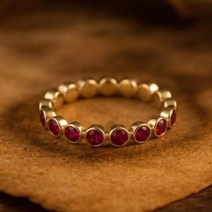 May include: A gold ring with a series of round, vibrant red gemstones set around the band. The ring is displayed on a textured, brown surface. The words "GRIFFANY ATELIER" are at the top of the image.