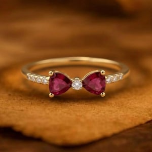 May include: A gold ring featuring a bow-shaped design with two red gemstones and a central white gemstone. The ring band is adorned with small white gemstones. The ring is set against a brown background.