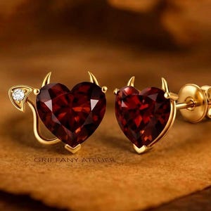 Könnte beinhalten: Ein Paar goldfarbene herzförmige Ohrstecker mit roten Edelsteinzentren. Jeder Ohrring hat kleine goldene Hörner und ein Schwanzdesign. Ein Ohrring hat einen kleinen Diamantakzent. Der Text "GRIFFANY ATELIER" ist sichtbar.