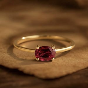 Puede incluir: Un anillo de oro con una gema roja oscura de forma ovalada. El anillo tiene una banda delgada y pulida, y la gema está sujeta por cuatro pequeñas garras. El anillo se presenta sobre un fondo cálido y neutro.