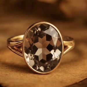 May include: A gold ring featuring a large, faceted, oval-cut gemstone with a smoky brown hue. The ring has a delicate, split-shank design and is set against a warm, neutral background.