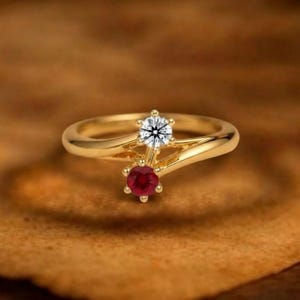 May include: A gold bypass ring featuring two round-cut stones set in six-prong settings. One stone is a clear, faceted white diamond, and the other is a deep red, faceted ruby. The band is smooth, polished yellow gold with a crossover design.