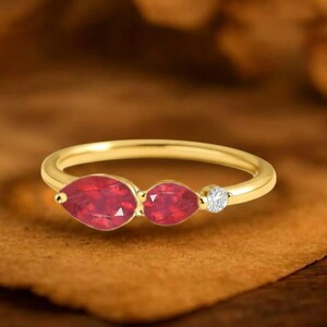 Peut inclure: Une bague en or ornée de deux pierres rouges en forme de poire et d'une petite pierre ronde et transparente. La bague est posée sur une surface marron. Le texte "GRIFFANY ATELIER" est en haut de l'image.