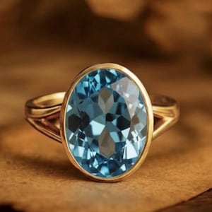 Puede incluir: Un anillo de oro con una gran piedra preciosa ovalada de color azul claro. La piedra preciosa está engastada en un bisel de oro, y el anillo tiene un diseño de caña dividida. El anillo se muestra sobre una superficie marrón.