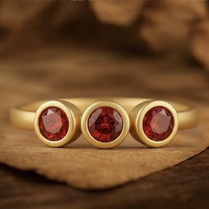 May include: A gold ring with three red gemstones. Each gemstone is round and set in a bezel. The ring's band is plain gold. The ring is shown on a textured, brown surface. The ring is from Griffany Atelier.