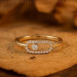 May include: A gold ring featuring an elongated oval frame set with small round diamonds. A single round diamond is positioned inside the oval frame, capable of sliding along the horizontal bar. The band is polished gold with a smooth finish.